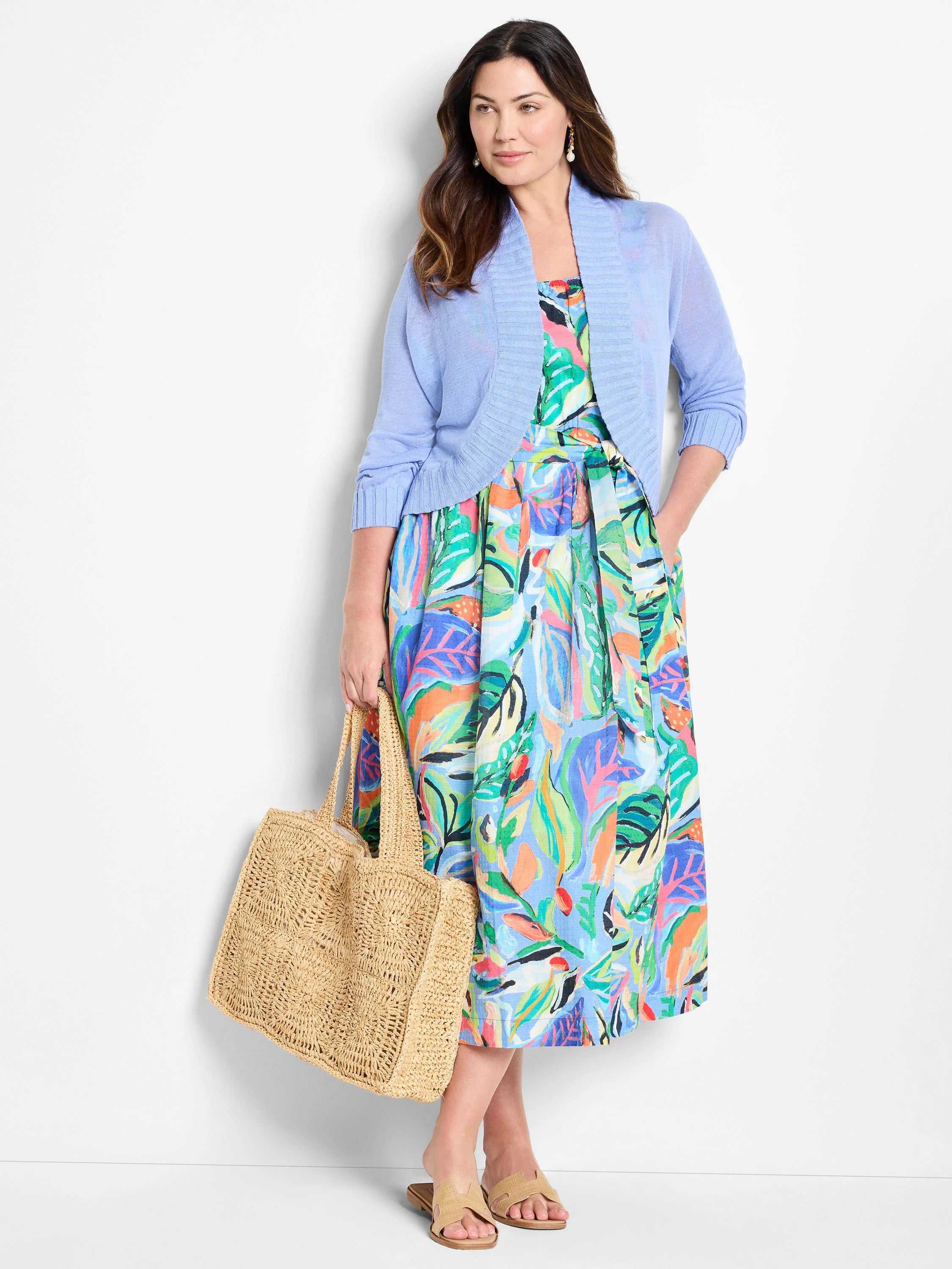 Model wears light blue cardigan over midi dress with vibrant tropical print in pinks, oranges, greens, and blues.  She...