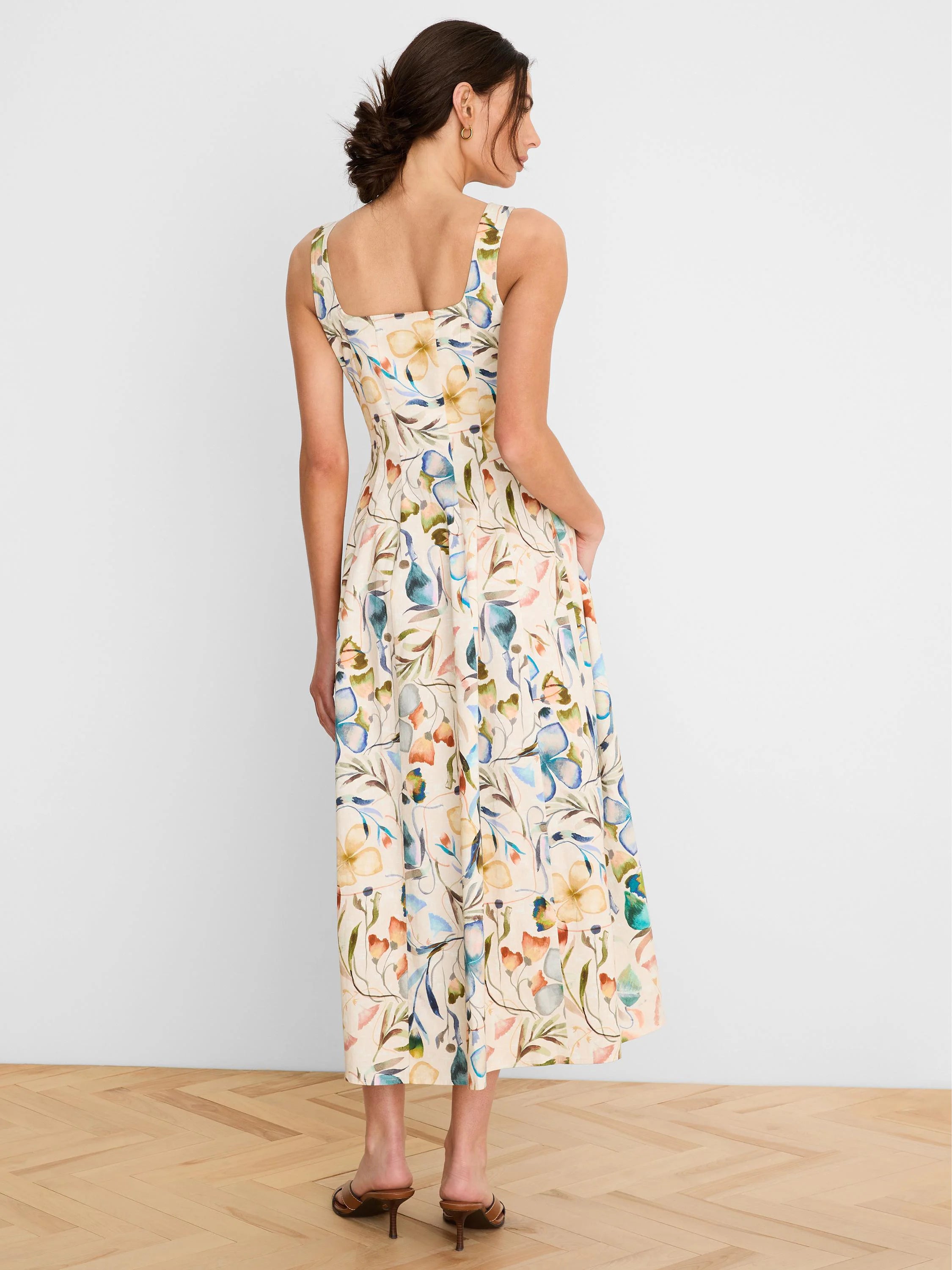 Sleeveless cream midi dress, model from back, hands in pockets. Features watercolor print: blue, green, yellow, and...