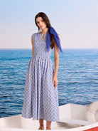 White sleeveless midi dress with blue embroidered tile pattern, smocked bodice, and flowy skirt. Blue scarf draped.