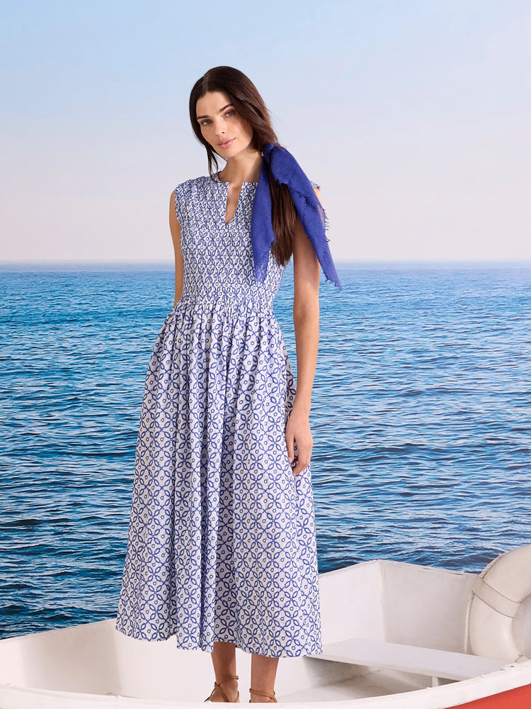 White sleeveless midi dress with blue embroidered tile pattern, smocked bodice, and flowy skirt. Blue scarf draped.