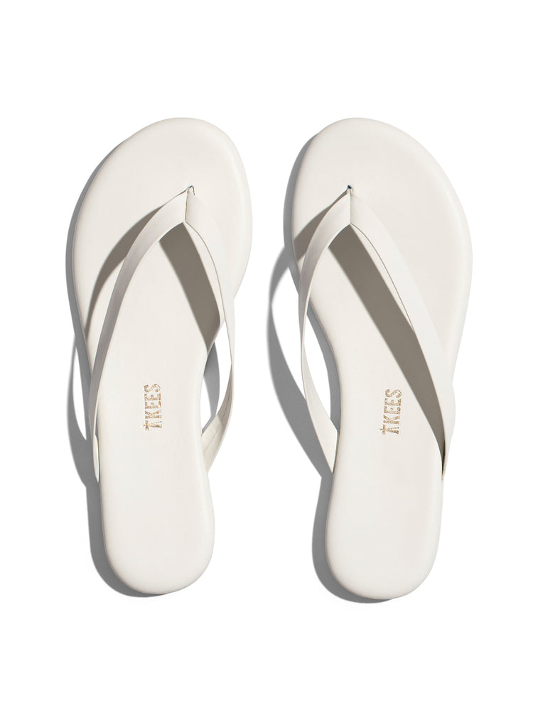 A pair of off-white leather flip-flops with thin straps and a flat sole exhibits a simple, minimalist design.