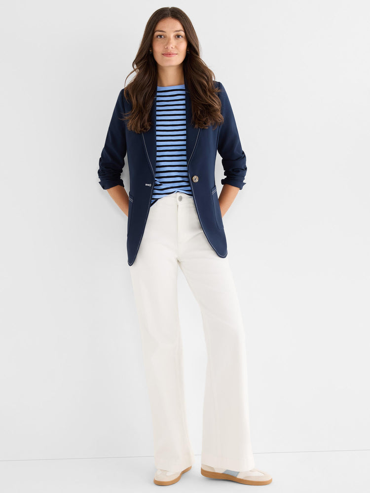 A model wears a navy fitted blazer featuring contrast white topstitching and rolled sleeves, styled open over a striped shirt.