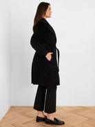 Black mid-length wrap coat, shown in profile, with wide lapels, a tie belt, and side pockets.