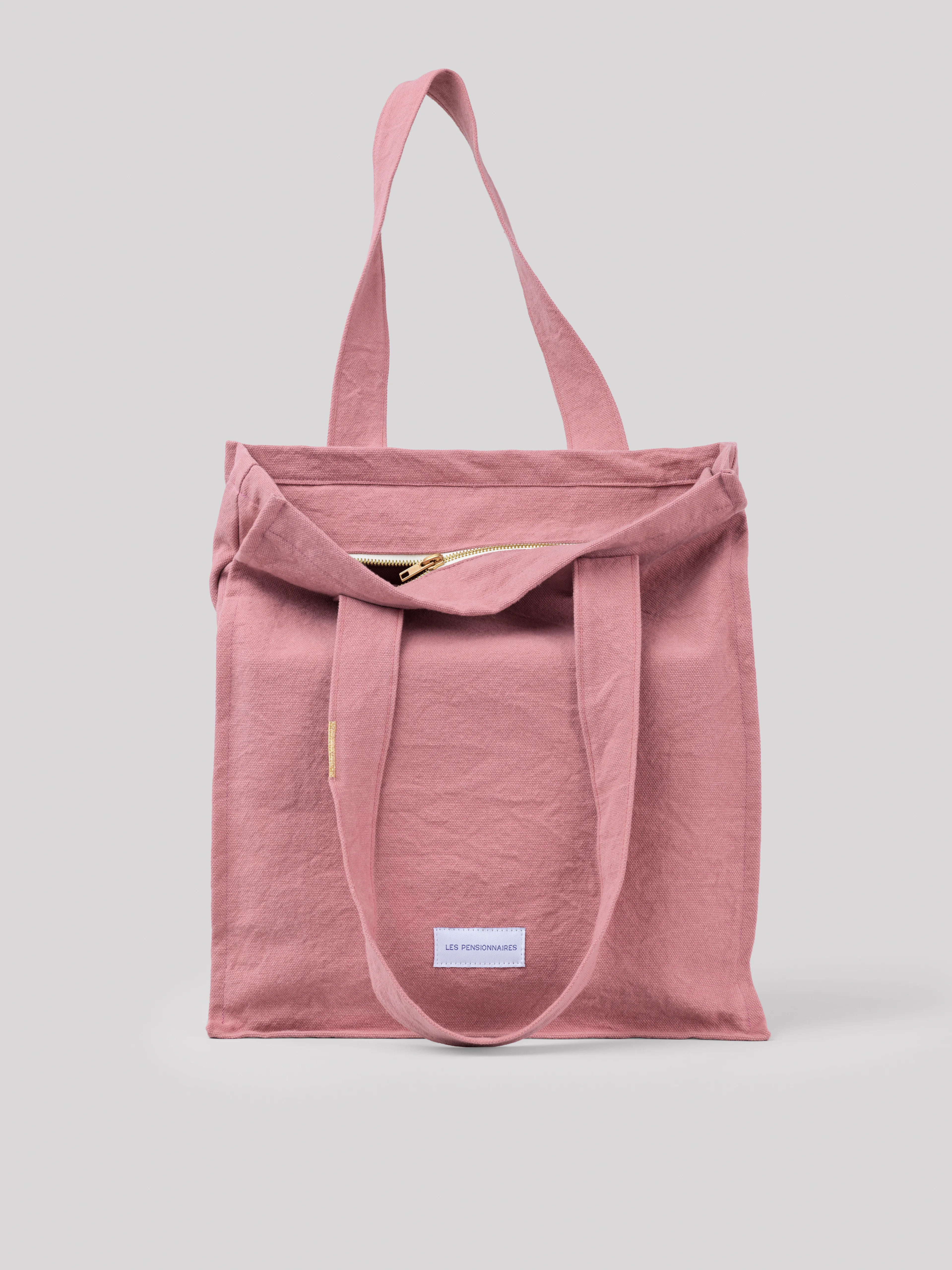 Pink cotton tote bag. It features a visible, partly unzipped gold zipper and two wide top handles. Small white label on...