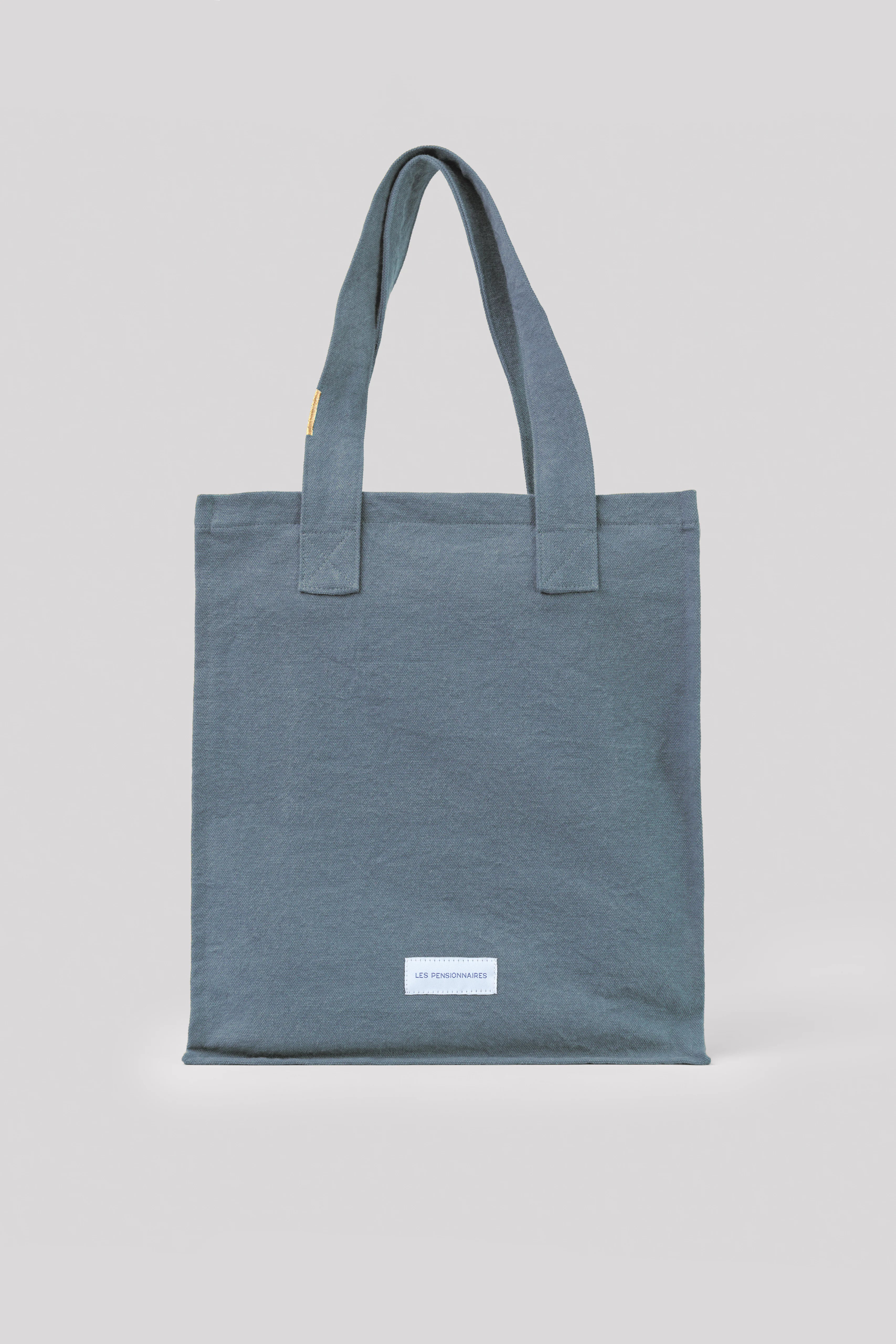 Slate blue cotton tote bag with two fabric handles and a white "LES PENSIONNAIRES" label on the front.