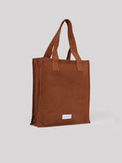 Rust cotton tote bag with two handles, a zippered top, and a small white brand label near the bottom.