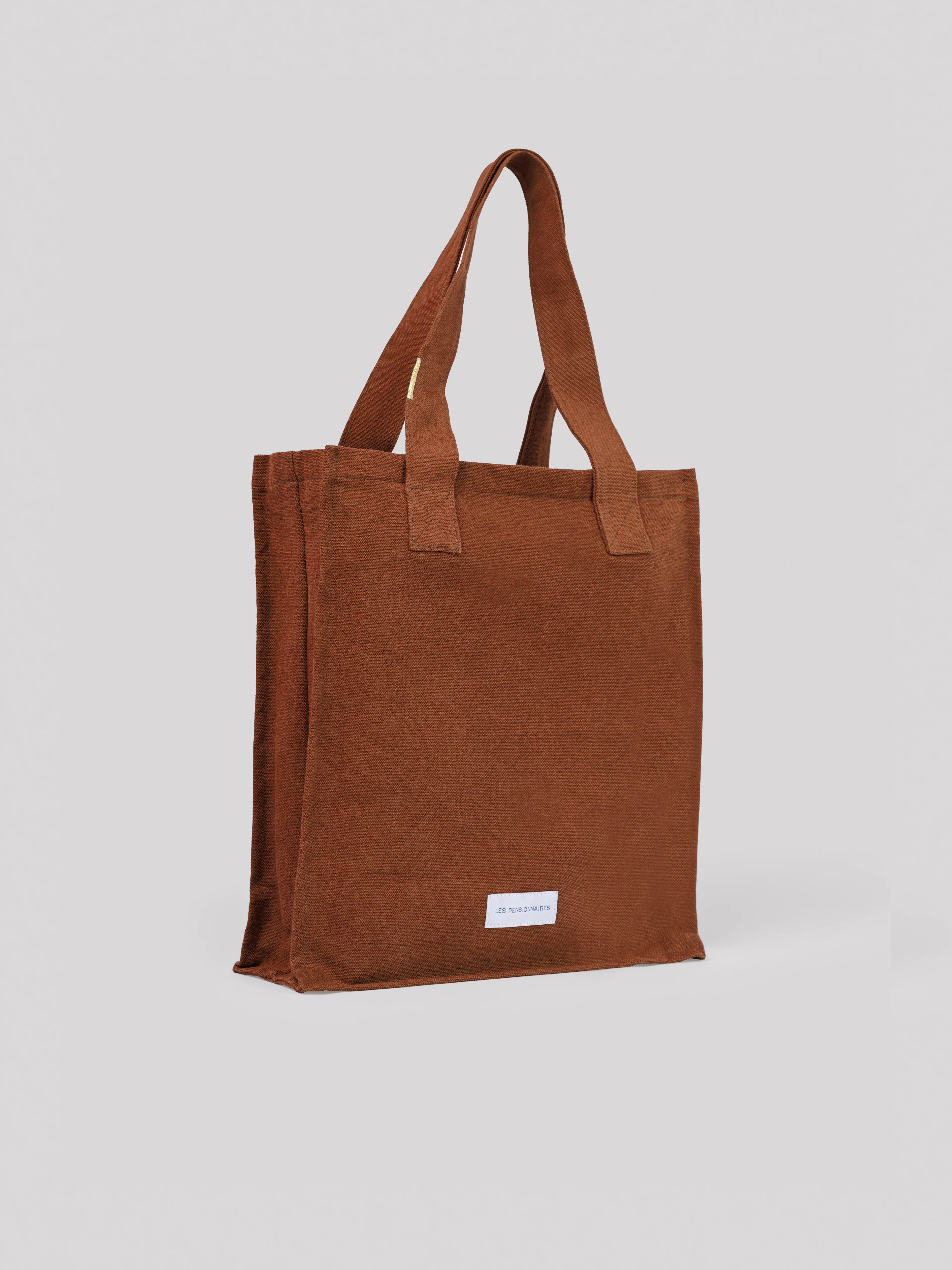 Rust cotton tote bag with two handles, a zippered top, and a small white brand label near the bottom.
