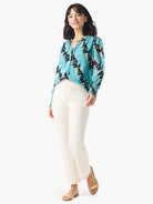 Teal top with dark & beige abstract print, long sleeves, and a V-neck.  Model also wears off-white pants and gold sandals.