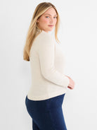 She wears a fitted beige and cream seed stitch mock neck sweater paired with dark wash jeans.