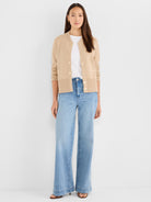 The model wears a fitted beige knit jacket featuring a crewneck, ribbed trim, and gold buttons over wide-leg denim.