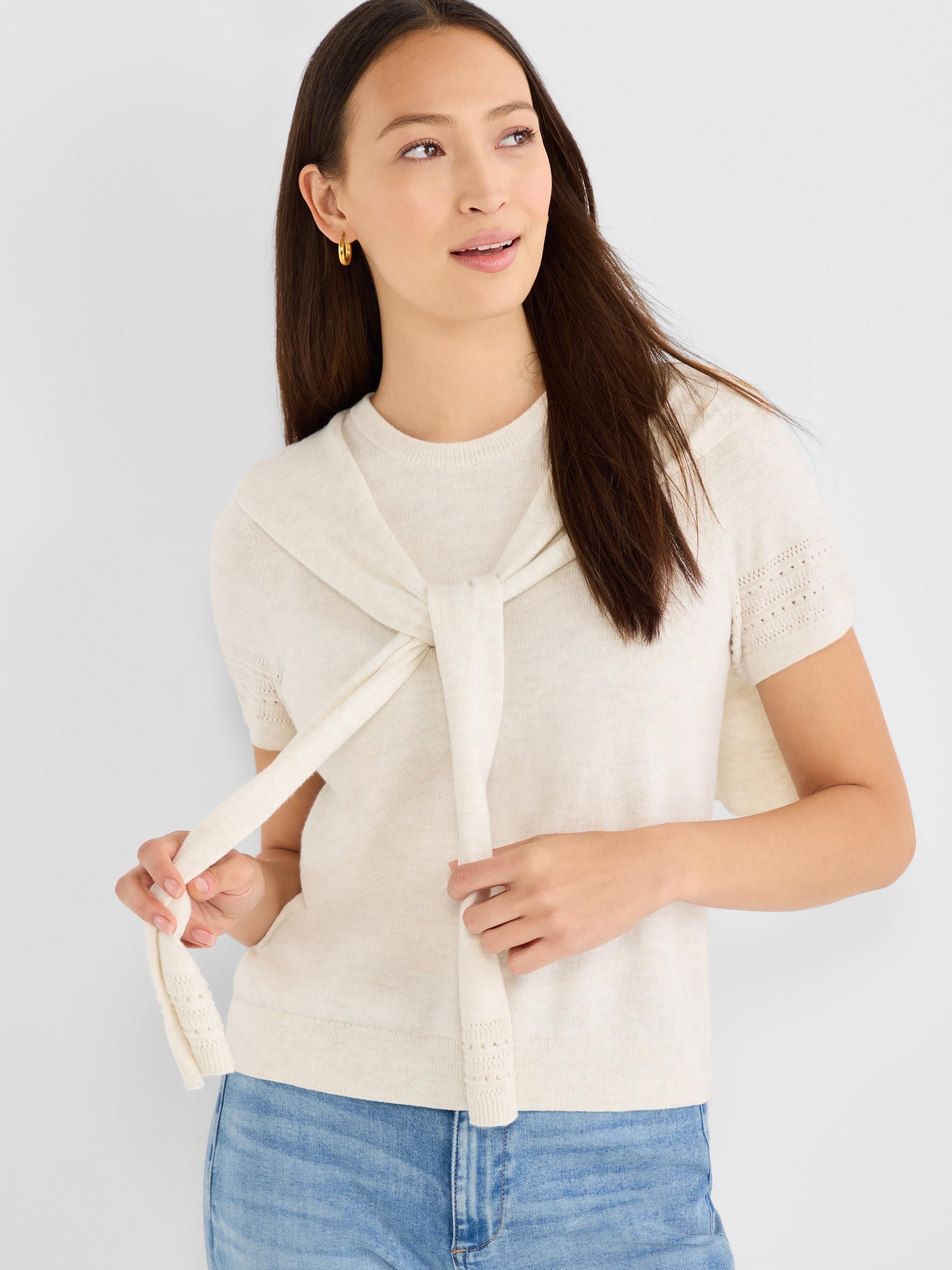 The model wears an oatmeal short-sleeve knit top with subtle eyelet sleeves, casually styled with a matching sweater tied.