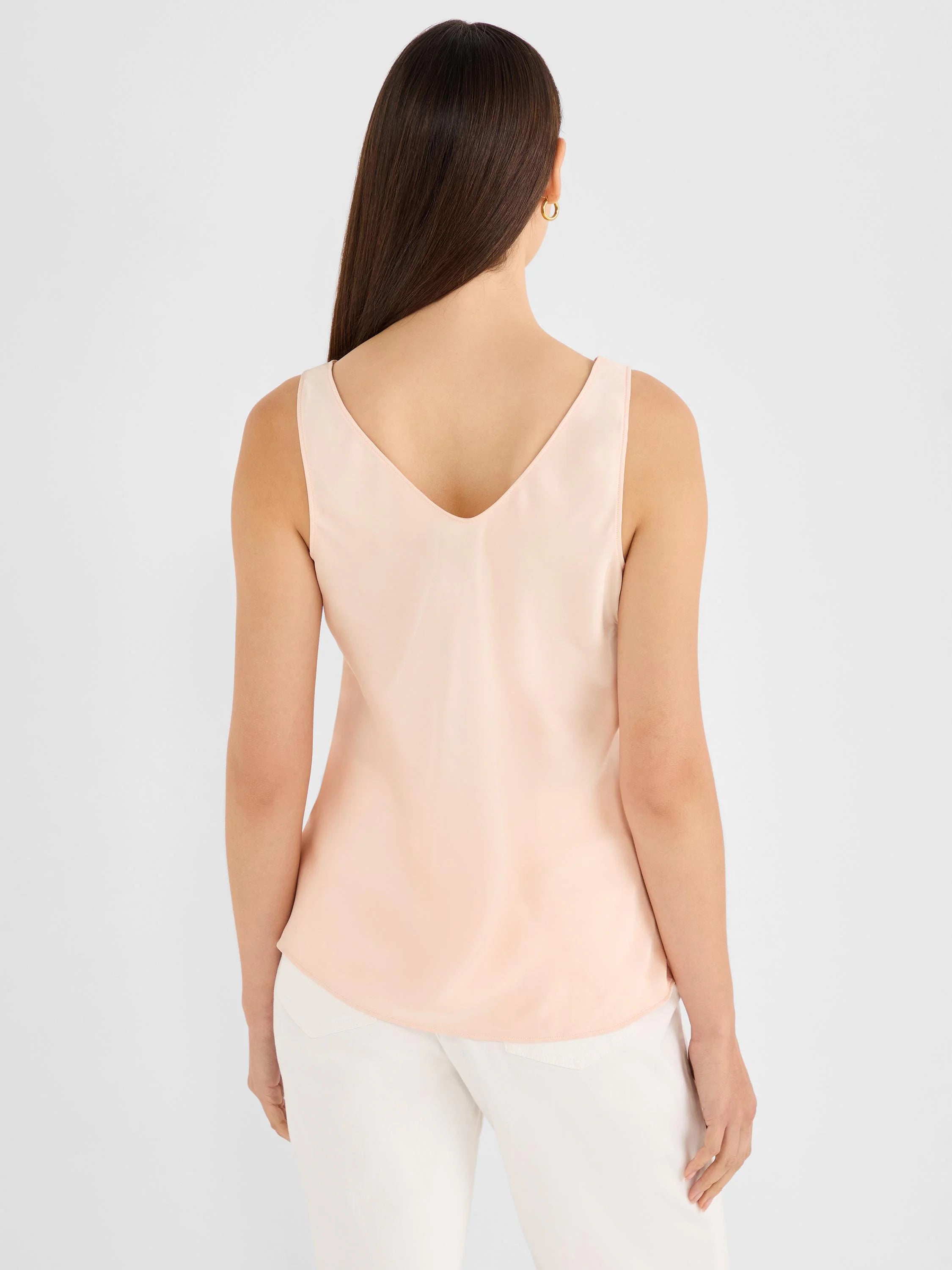 Back of a light peach woven sleeveless tank with a V-neck. Model has long brown hair, wears white pants.