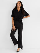 She wears a relaxed black short-sleeve collared top paired with high-waisted tailored black bootcut pants.