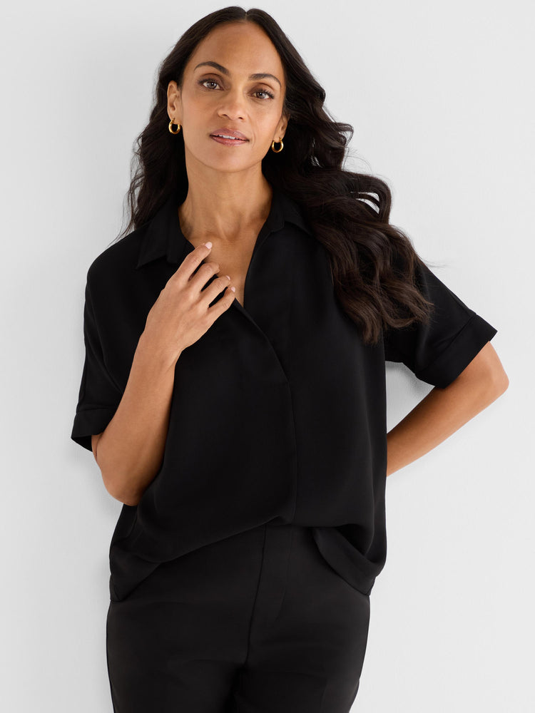 A black short-sleeve draped blouse with a relaxed fit and open collar is styled professionally with black trousers.