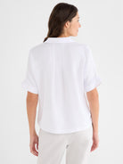 Model wears a relaxed white collared blouse with cuffed short sleeves and a structured back seam.