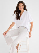 The model wears a relaxed white short-sleeve blouse and light taupe wide-leg trousers featuring gold button accents.