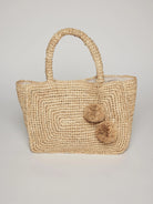 Natural woven straw tote bag with two top handles and two fluffy straw pom-poms on the front.