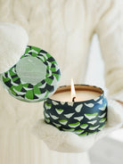 Round, dark teal tin candle with green & white abstract pattern, lit flame, lid ajar, held by gloved hands.