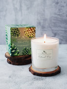 Lit white candle in clear glass jar on wood slice coaster, green box with pinecone design behind.
