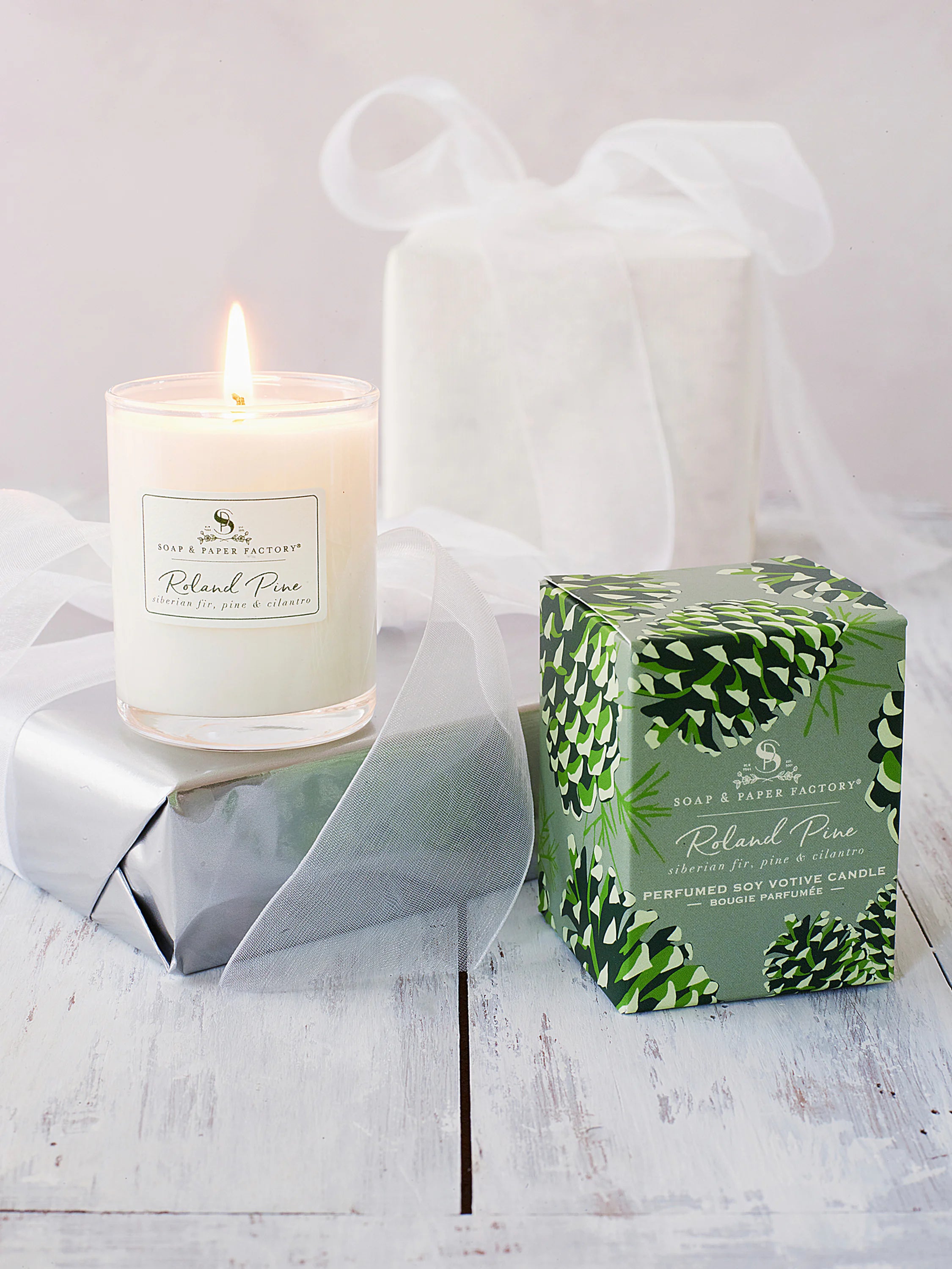 White votive candle, lit in clear glass. Sage green box with dark and light green pinecone patterns.