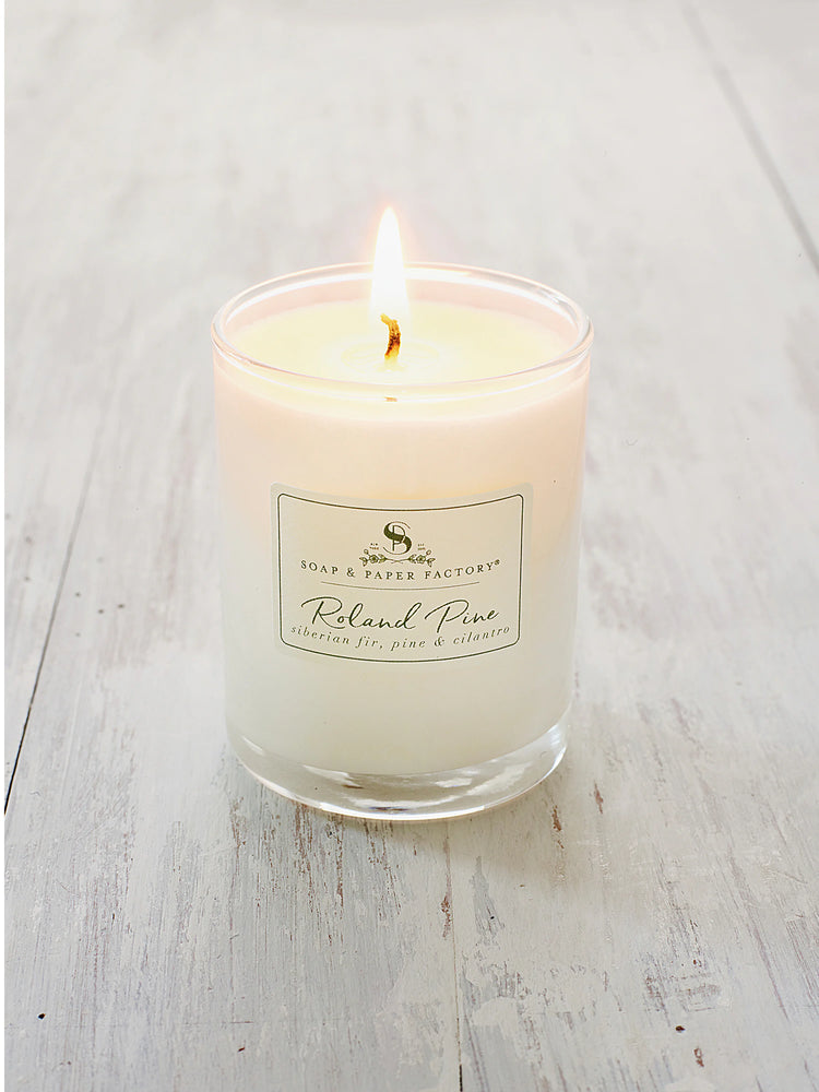 Lit votive candle with creamy white wax in a clear glass holder, featuring a 'Roland Pine' label, on a light, distressed...