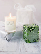 A lit white candle in a clear glass votive holder, next to its sage green box with a pinecone pattern.