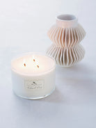 Off-white soy candle in clear glass, three lit wicks.  Small off-white label.  Beside a textured off-white vase.