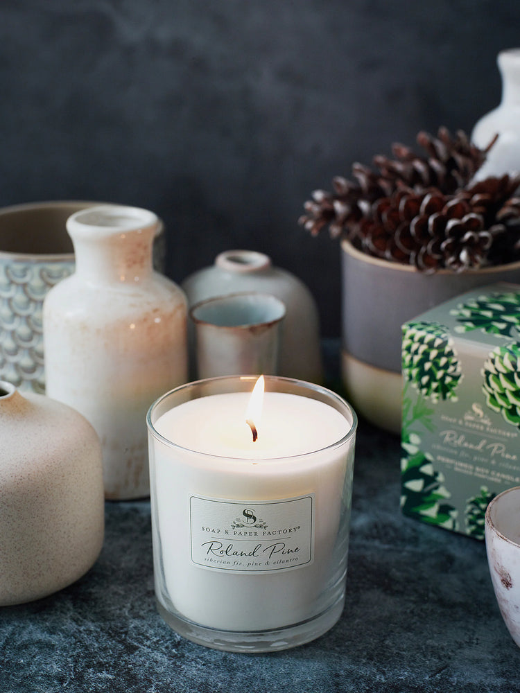 Off-white soy candle in clear glass, burning, on dark surface.  Label shows product name & scent notes.