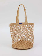 Tan woven jute tote bag with an open mesh body, braided straps, and a small blue label at the top.