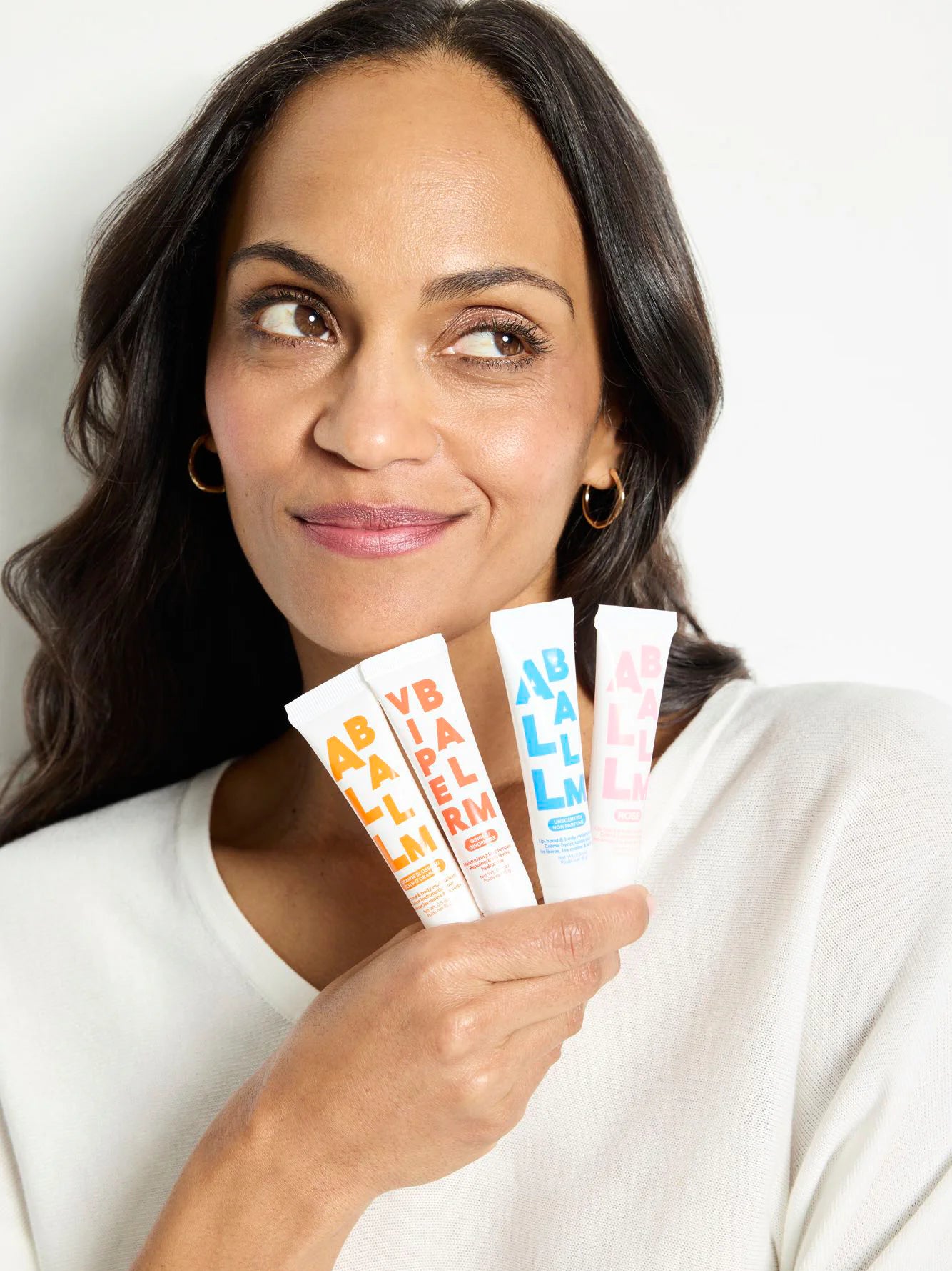 Woman holds a white Zizia Rose All Balm tube with pink 'BALM' text, plus orange and blue variants.
