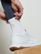 White ribbed sock with subtle tan V-symbols. Worn on an ankle with dark leggings and white shoe.