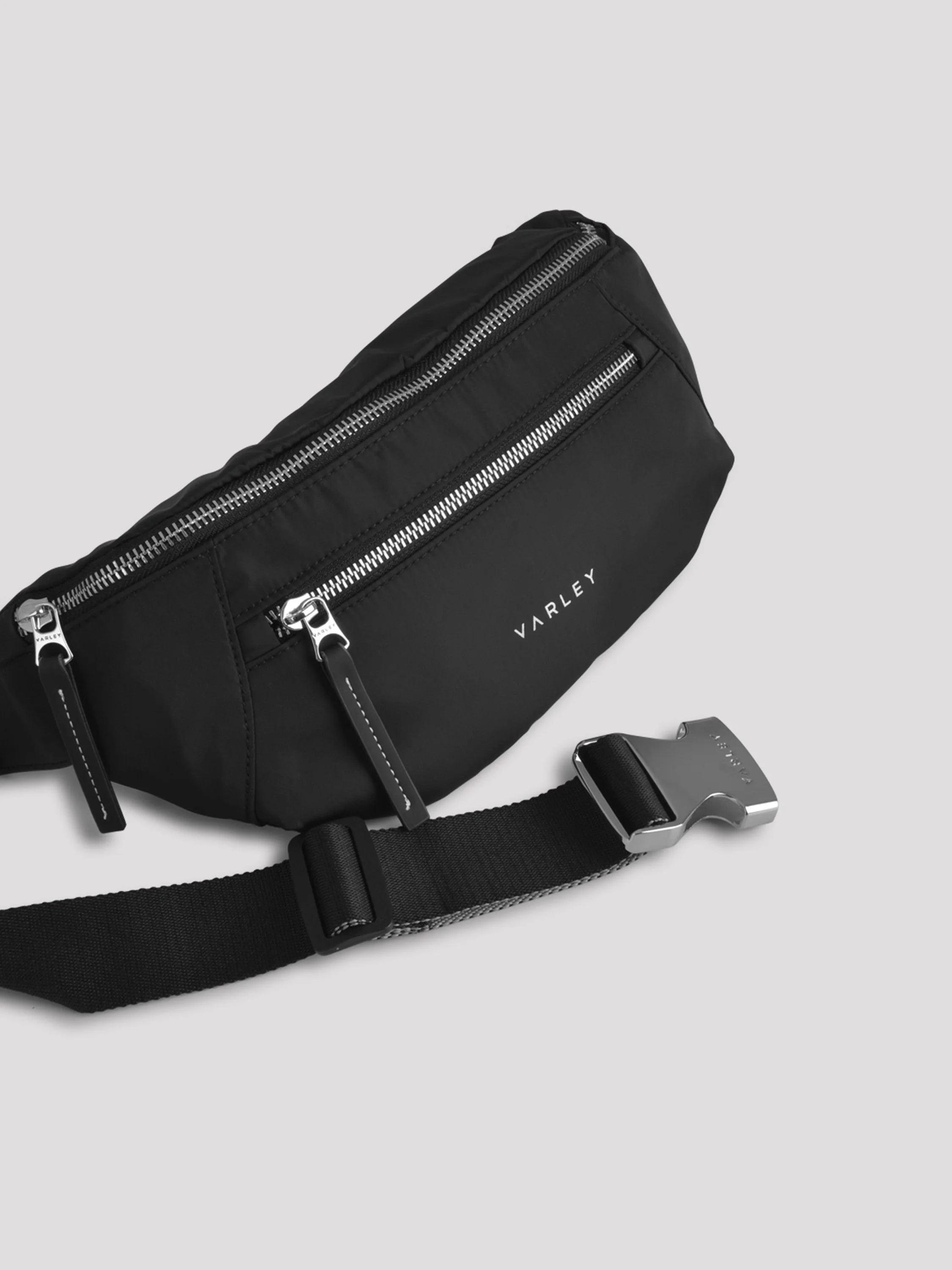 Black fabric belt bag with two silver zippers, a black strap, and a silver buckle. White 'VARLEY' logo on front.