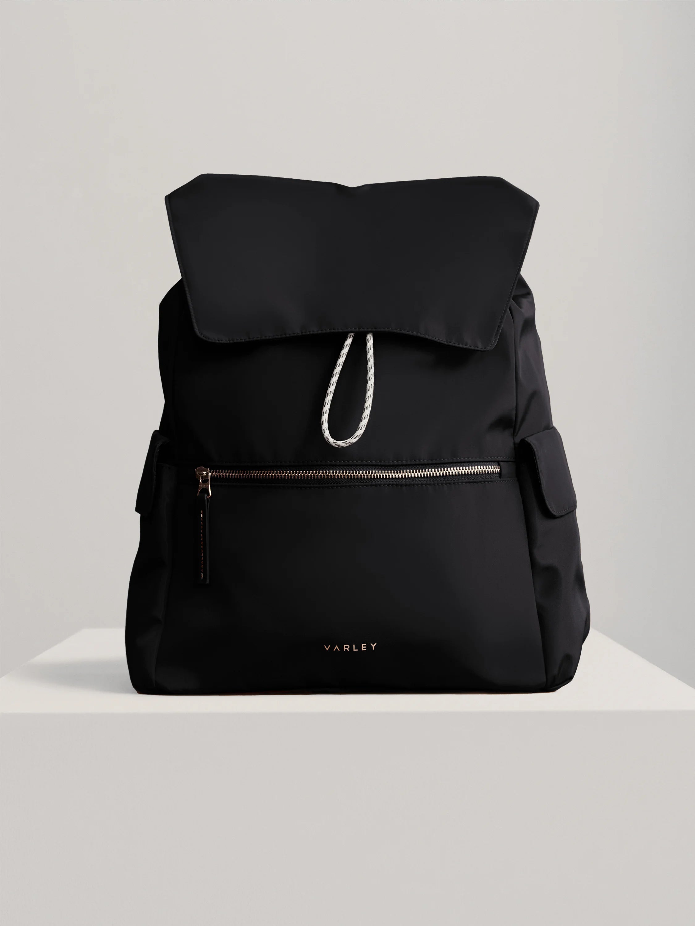 Black backpack with a top flap, white drawstring, gold front zipper, and gold Varley logo.