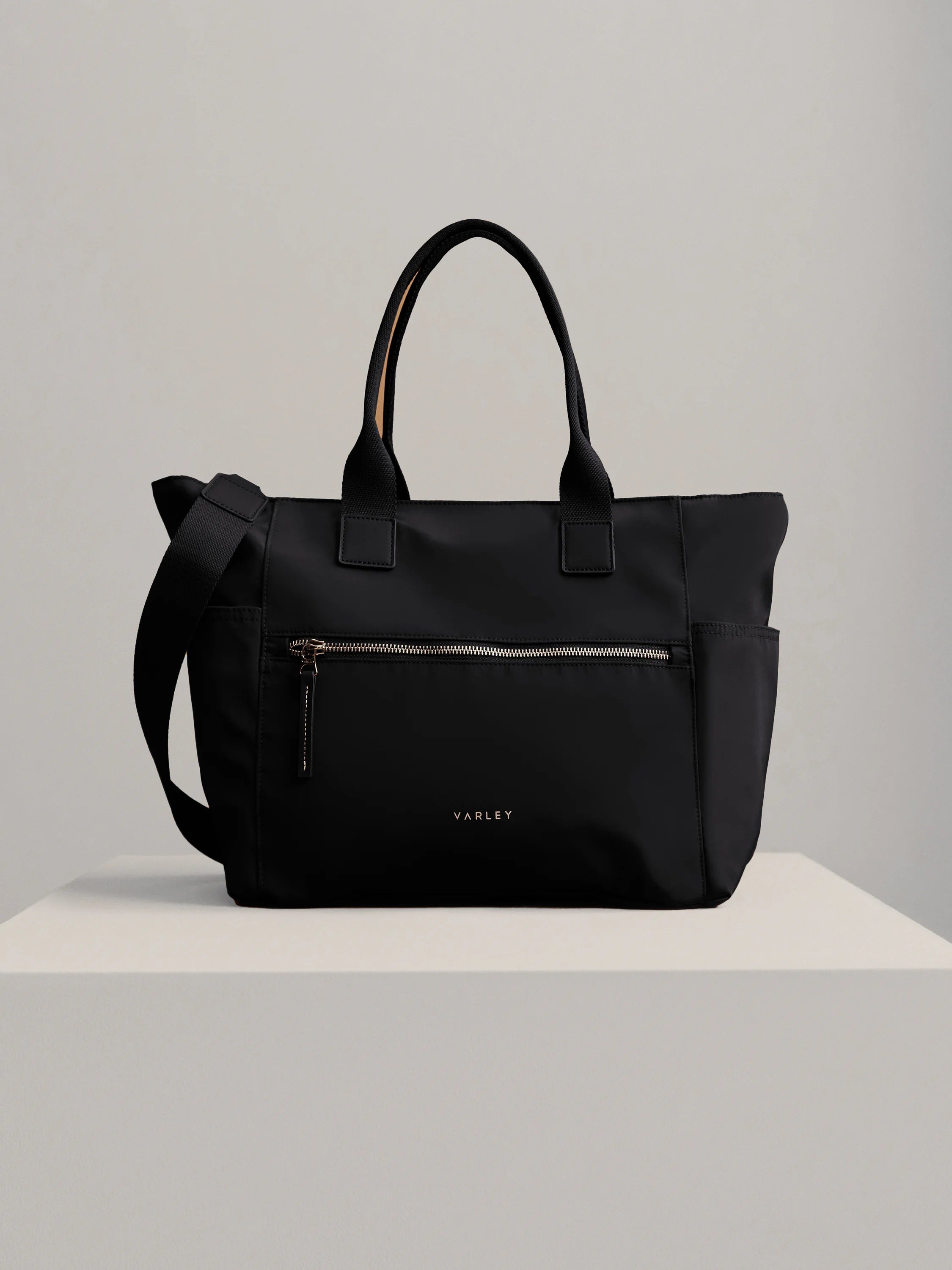 Matte black Varley Kelso crossbody bag with top handles, adjustable strap, front zip pocket, side pockets, and metallic logo.