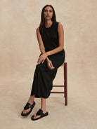 Black sleeveless maxi dress with a round neck and gathered waist, worn by a model seated.