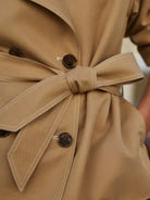 Camel short trench coat with a bow-tied belt, dark buttons, and white contrast stitching detail.