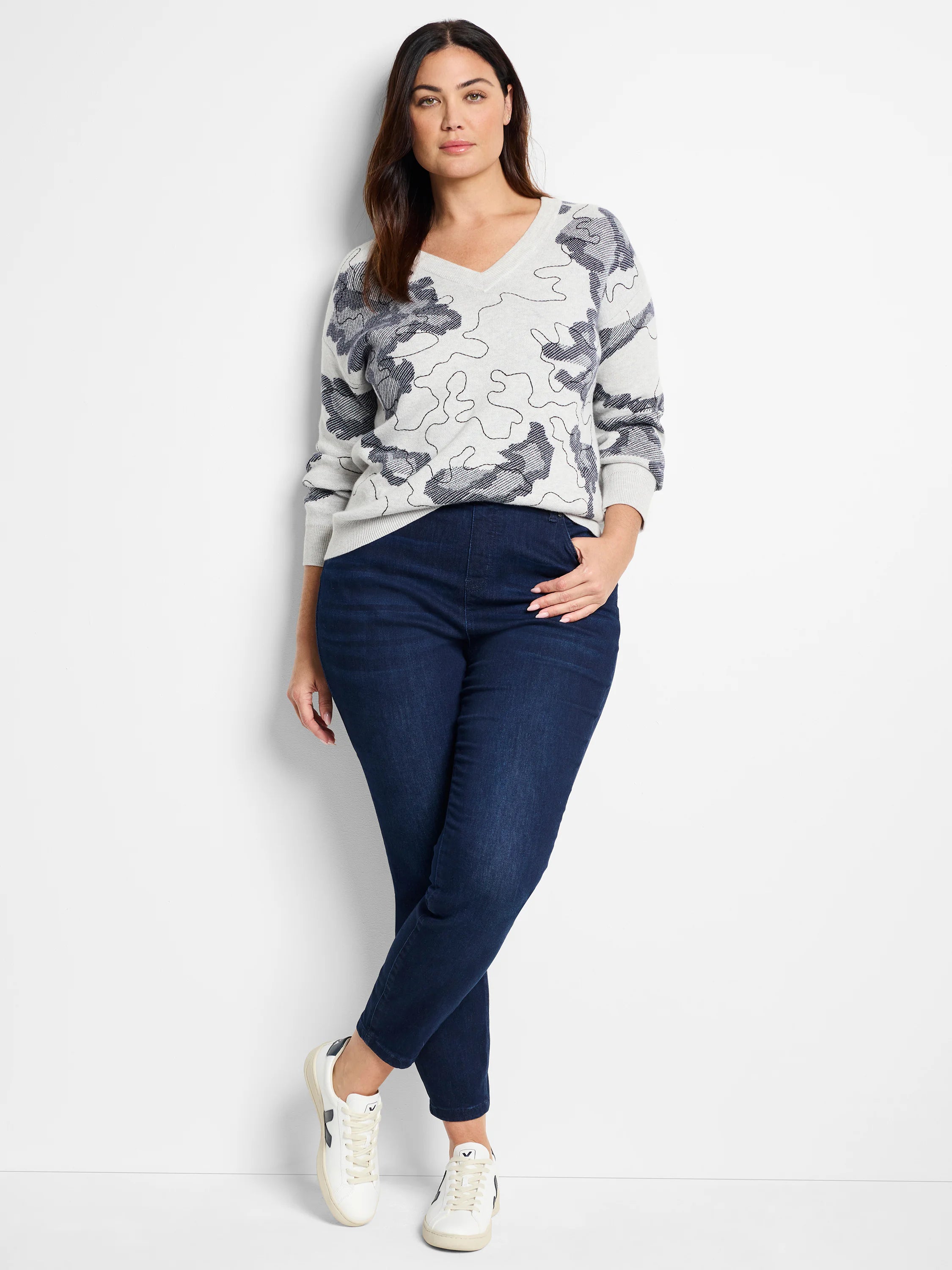 Light gray V-neck sweater with dark gray abstract pattern.  Model also wears dark wash jeans and white sneakers.