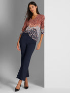 She wears a dotted V-neck blouse in warm tones over fitted, navy, cropped ankle pants.