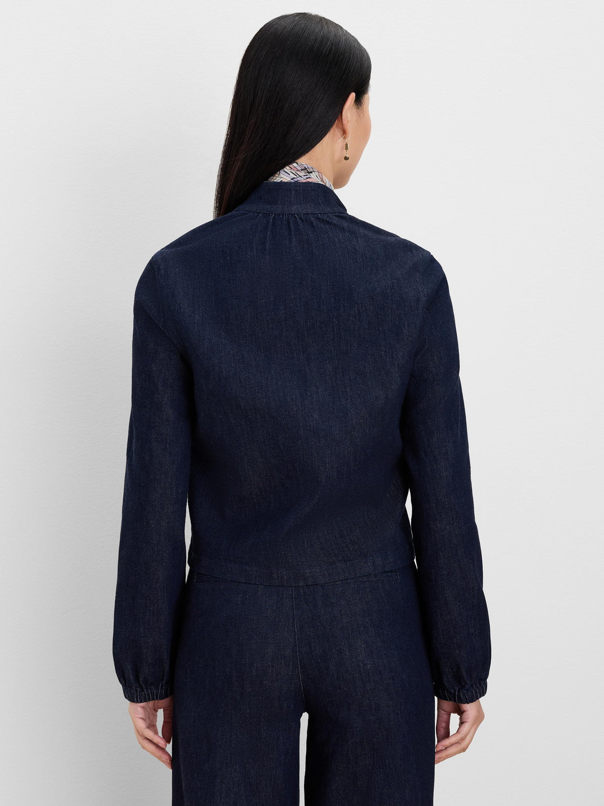 Dark indigo denim jacket, back view. Features a band collar and long sleeves with gathered elastic cuffs.