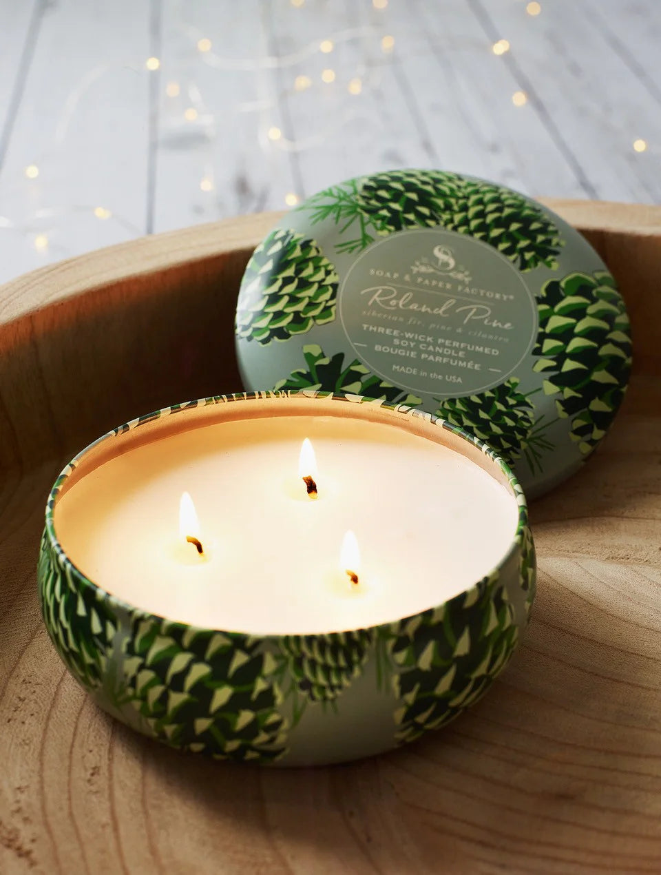 Round light green tin candle with dark green pinecone pattern, three lit wicks, and matching lid beside it.